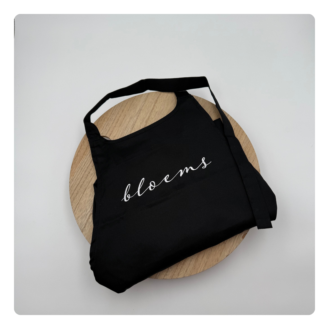 Zwart keukenschort met geborduurde tekst “Bloems” op de borst – gepersonaliseerd door Mbroidery